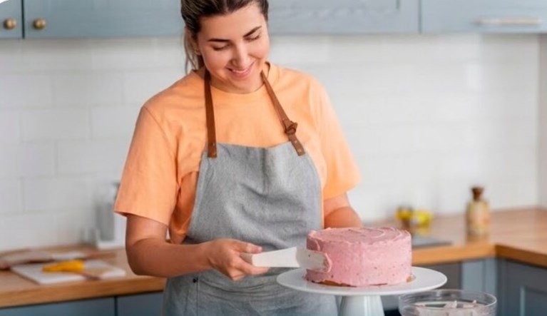 Woman in an orange shirt and gray apron frosting a pink cake on a white cake stand in a bright kitchen