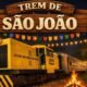 Wooden sign reading Trem de São João above a festive night scene with a yellow train car string lights and market stalls in the background