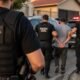 Civil police officers detain a man on a residential street as police cars with flashing lights are visible in the background