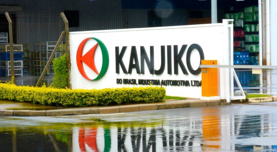 Industrial facility entrance with a white sign reading KANJIKO and a red-green circular logo; wet pavement reflects the sign.