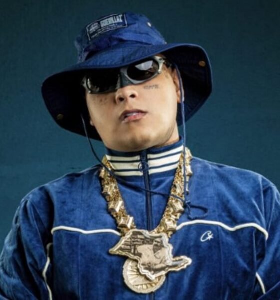 Person in a blue velour tracksuit with white piping blue bucket hat sunglasses and heavy gold chains against a dark teal background