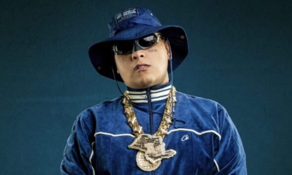 Person in a blue velour tracksuit with white piping blue bucket hat sunglasses and heavy gold chains against a dark teal background