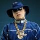 Person in a blue velour tracksuit with white piping blue bucket hat sunglasses and heavy gold chains against a dark teal background