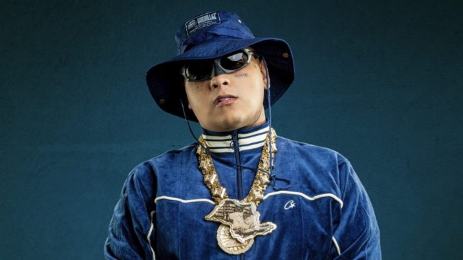 Person in a blue velour tracksuit with white piping blue bucket hat sunglasses and heavy gold chains against a dark teal background