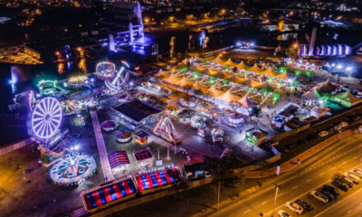 Aerial night view of a waterfront carnival with illuminated tents, rides, and a large Ferris wheel.