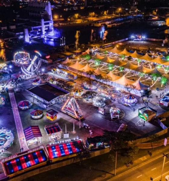 Aerial night view of a waterfront carnival with illuminated tents rides and a large Ferris wheel