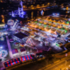 Aerial night view of a waterfront carnival with illuminated tents rides and a large Ferris wheel
