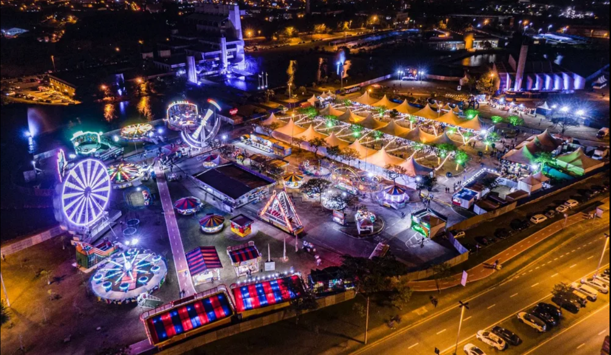 Aerial night view of a waterfront carnival with illuminated tents rides and a large Ferris wheel