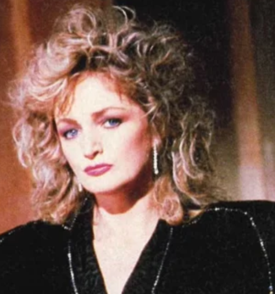 Portrait of a woman with voluminous blonde curly hair wearing a black velvet jacket and dangling earrings looking at the camera