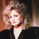 Portrait of a woman with voluminous blonde curly hair wearing a black velvet jacket and dangling earrings looking at the camera