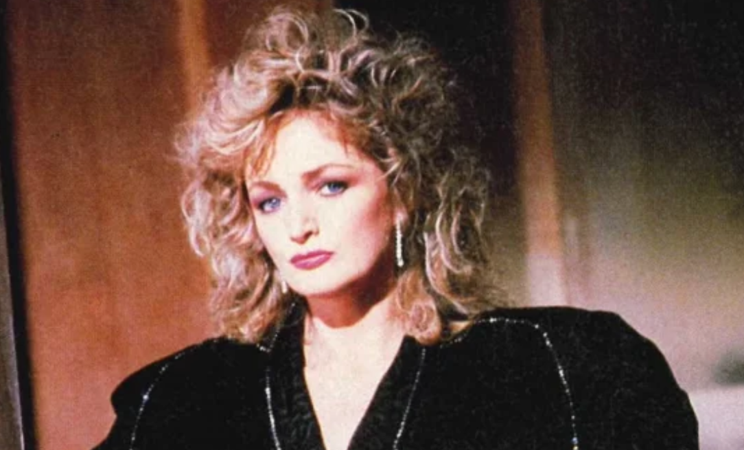 Portrait of a woman with voluminous blonde curly hair wearing a black velvet jacket and dangling earrings looking at the camera