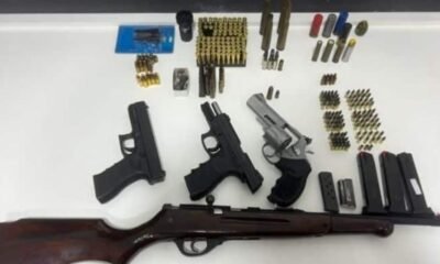 Three handguns and a long rifle laid out on a white table with numerous magazines ammo boxes and rounds arranged nearby