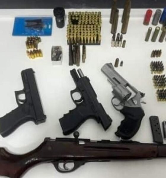 Three handguns and a long rifle laid out on a white table with numerous magazines ammo boxes and rounds arranged nearby