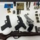 Three handguns and a long rifle laid out on a white table with numerous magazines ammo boxes and rounds arranged nearby