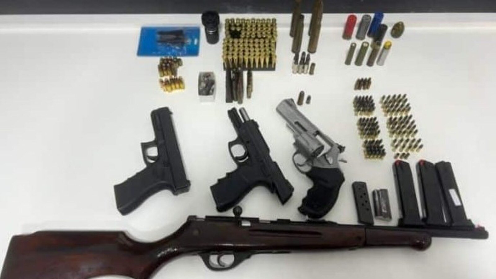Three handguns and a long rifle laid out on a white table with numerous magazines ammo boxes and rounds arranged nearby