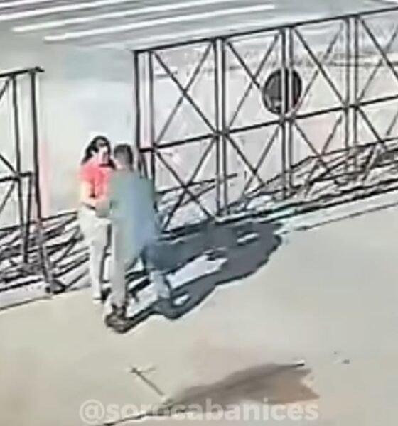 Two people standing close together by a metal lattice fence along a sidewalk outside a building one in a pink shirt
