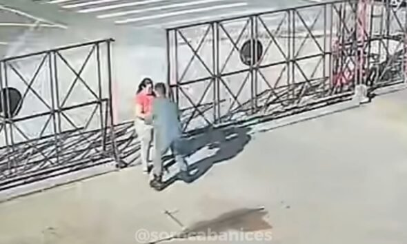 Two people standing close together by a metal lattice fence along a sidewalk outside a building one in a pink shirt