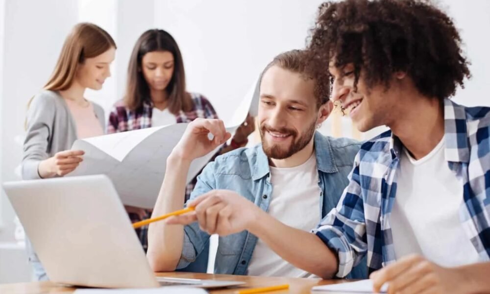 Group of colleagues collaborating around a laptop, two in the foreground smiling as they discuss with a pencil and plans nearby.