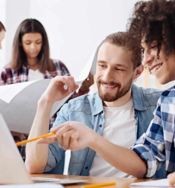 Group of colleagues collaborating around a laptop two in the foreground smiling as they discuss with a pencil and plans nearby