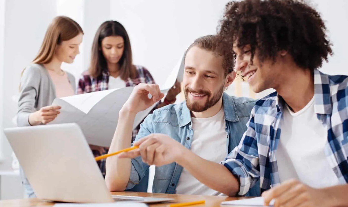 Group of colleagues collaborating around a laptop two in the foreground smiling as they discuss with a pencil and plans nearby