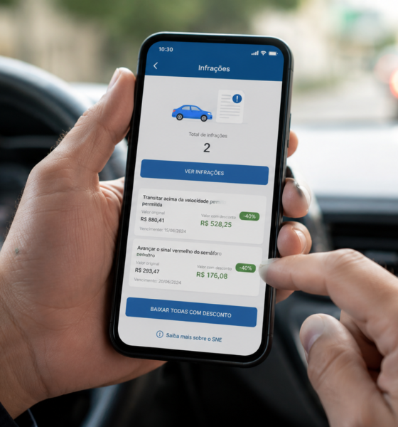Pessoa no carro segura o celular exibindo um app de infrações de trânsito com o total de  infrações na tela