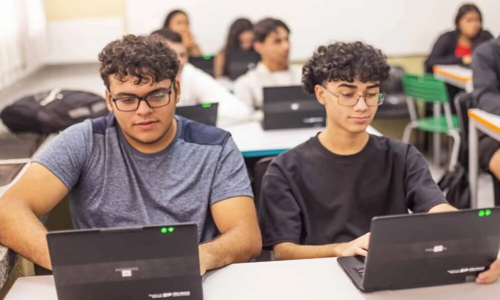 Two students sit at a classroom table working on laptops, focused on their screens as classmates learn in the background