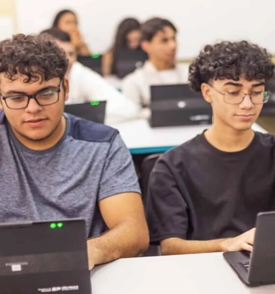 Two students sit at a classroom table working on laptops focused on their screens as classmates learn in the background