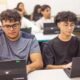 Two students sit at a classroom table working on laptops focused on their screens as classmates learn in the background