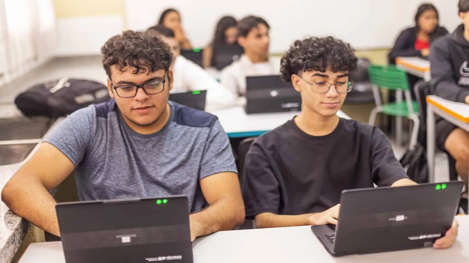 Two students sit at a classroom table working on laptops focused on their screens as classmates learn in the background