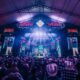 Large indoor concert stage with bright neon lights and a crowd facing the performers under a peaked roof festival tent