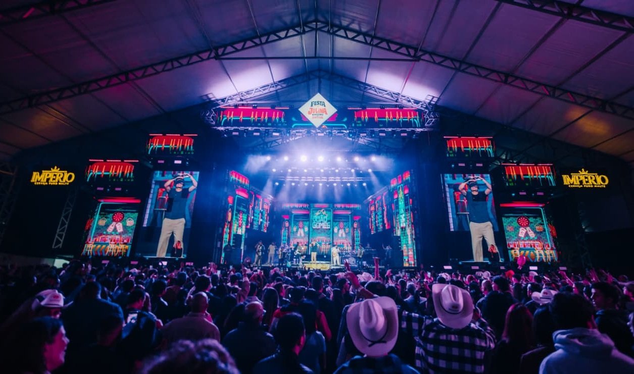 Large indoor concert stage with bright neon lights and a crowd facing the performers under a peaked roof festival tent