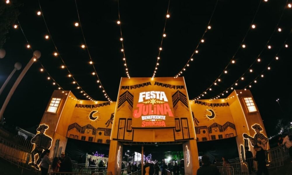 Night view of a Festa Julina entrance with yellow arches, string lights, and a crowd gathering under the festival setup, at night.