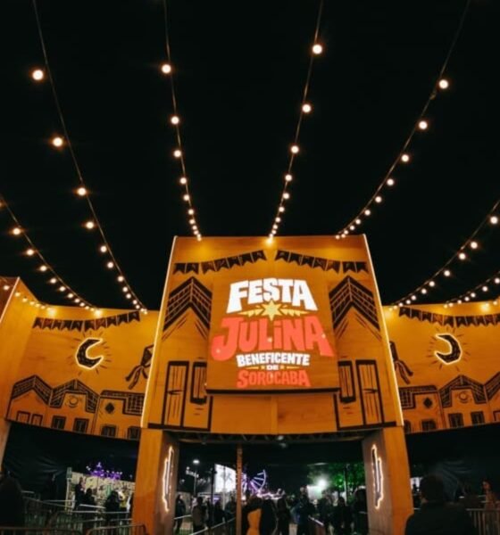 Night view of a Festa Julina entrance with yellow arches, string lights, and a crowd gathering under the festival setup, at night.