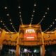 Night view of a Festa Julina entrance with yellow arches string lights and a crowd gathering under the festival setup at night