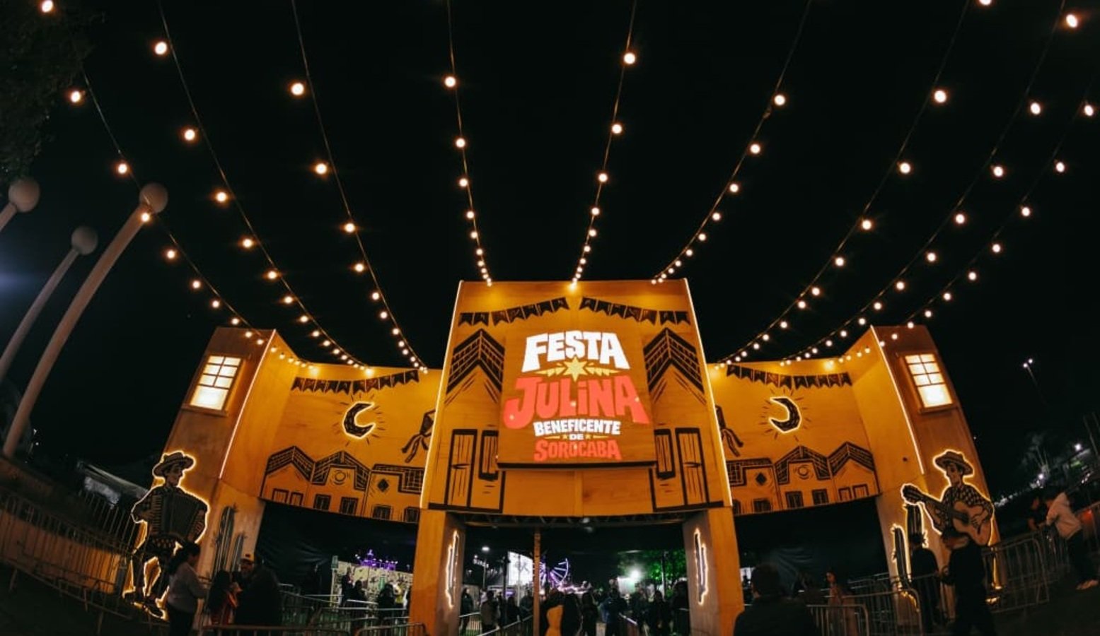 Night view of a Festa Julina entrance with yellow arches string lights and a crowd gathering under the festival setup at night