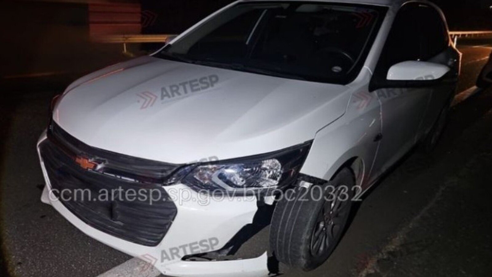 White Chevrolet sedan at night with front bumper damaged and hanging parked on a dark road edge