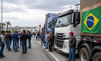 Group of truck drivers standing on a highway beside parked semi trucks with a Brazil flag on a trailer in the background