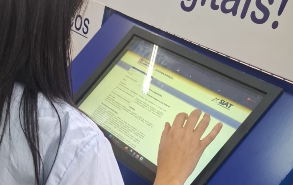 Person with long dark hair in a white shirt uses a large touchscreen kiosk to view a form SIAT logo visible on the screen area