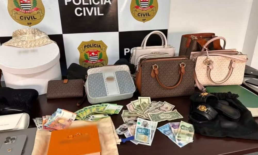 Seized collection of luxury handbags, wallets, and cash laid out on a table by Polícia Civil São Paulo backdrop