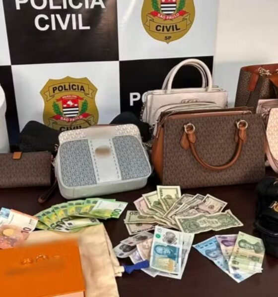 Seized collection of luxury handbags wallets and cash laid out on a table by Polícia Civil São Paulo backdrop