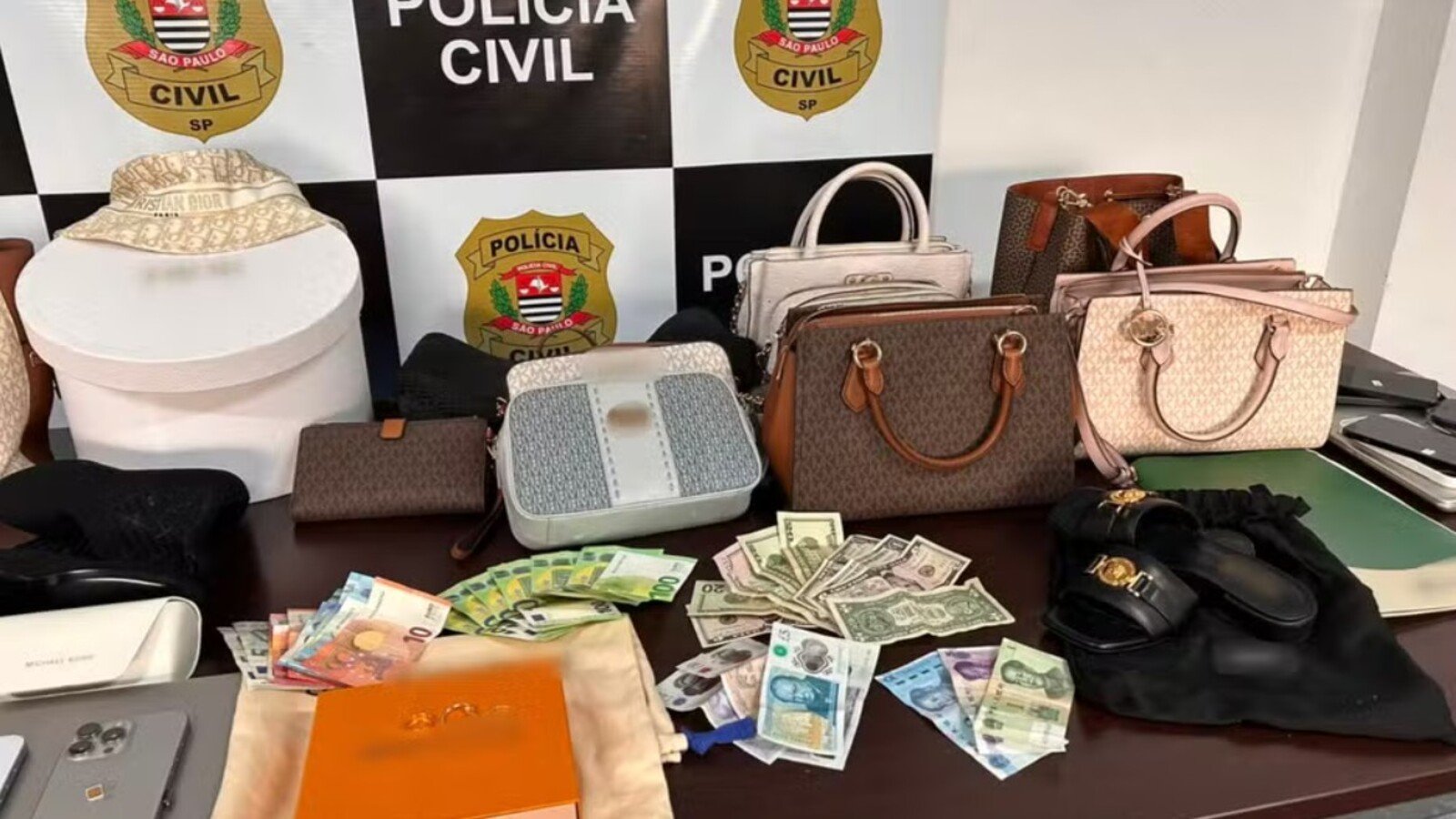 Seized collection of luxury handbags wallets and cash laid out on a table by Polícia Civil São Paulo backdrop