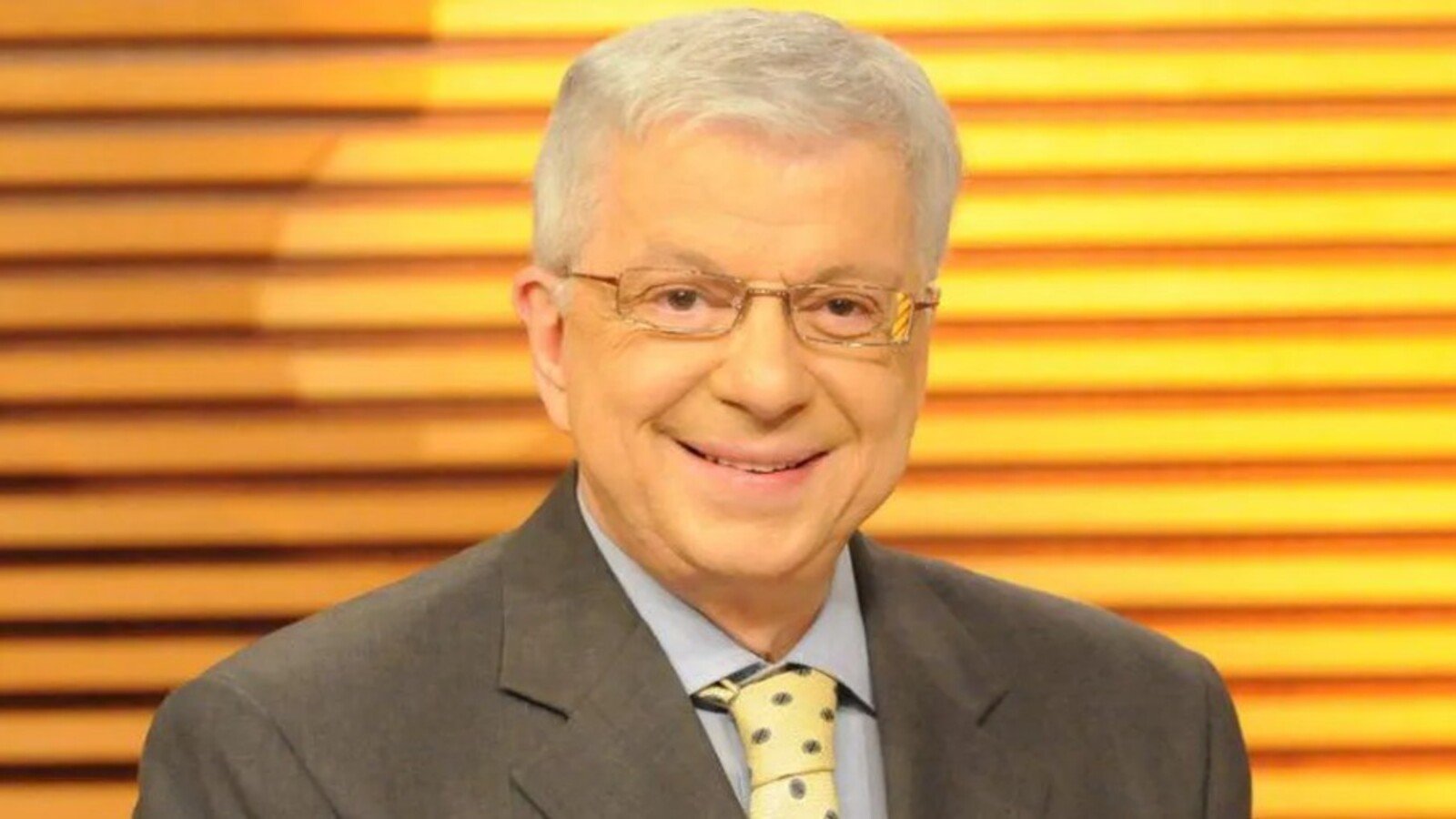 Smiling middle aged man in a gray suit and yellow polka dotted tie wearing glasses against a warm wooden slat background