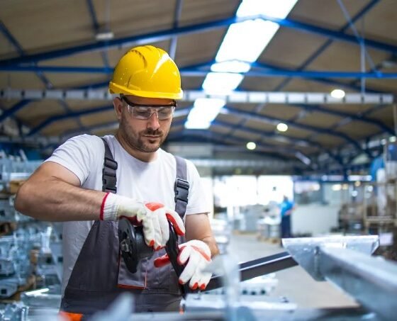 Worker in a yellow hard hat safety glasses and gloves cuts a metal beam with a grinder in a busy warehouse