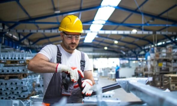 Worker in a yellow hard hat safety glasses and gloves cuts a metal beam with a grinder in a busy warehouse