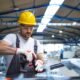Worker in a yellow hard hat safety glasses and gloves cuts a metal beam with a grinder in a busy warehouse