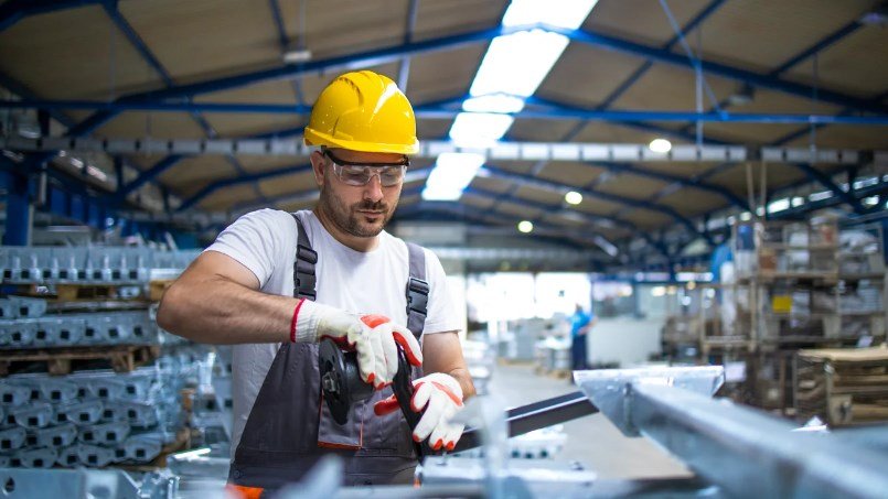 Worker in a yellow hard hat safety glasses and gloves cuts a metal beam with a grinder in a busy warehouse
