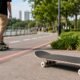 Empty skateboard lying on a paved path beside a grassy park with a person riding a longboard in the background by the water fence