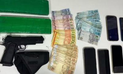 Assorted items on a table two large green wrapped bundles a black handgun with a holster and stacks of Brazilian real banknotes next to multiple mobile phones