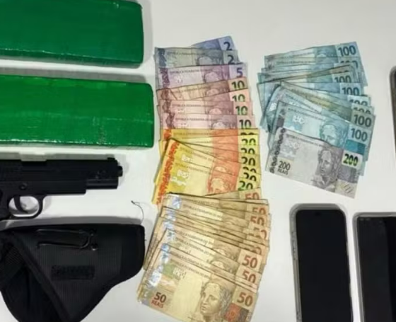 Assorted items on a table two large green wrapped bundles a black handgun with a holster and stacks of Brazilian real banknotes next to multiple mobile phones