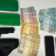 Assorted items on a table two large green wrapped bundles a black handgun with a holster and stacks of Brazilian real banknotes next to multiple mobile phones